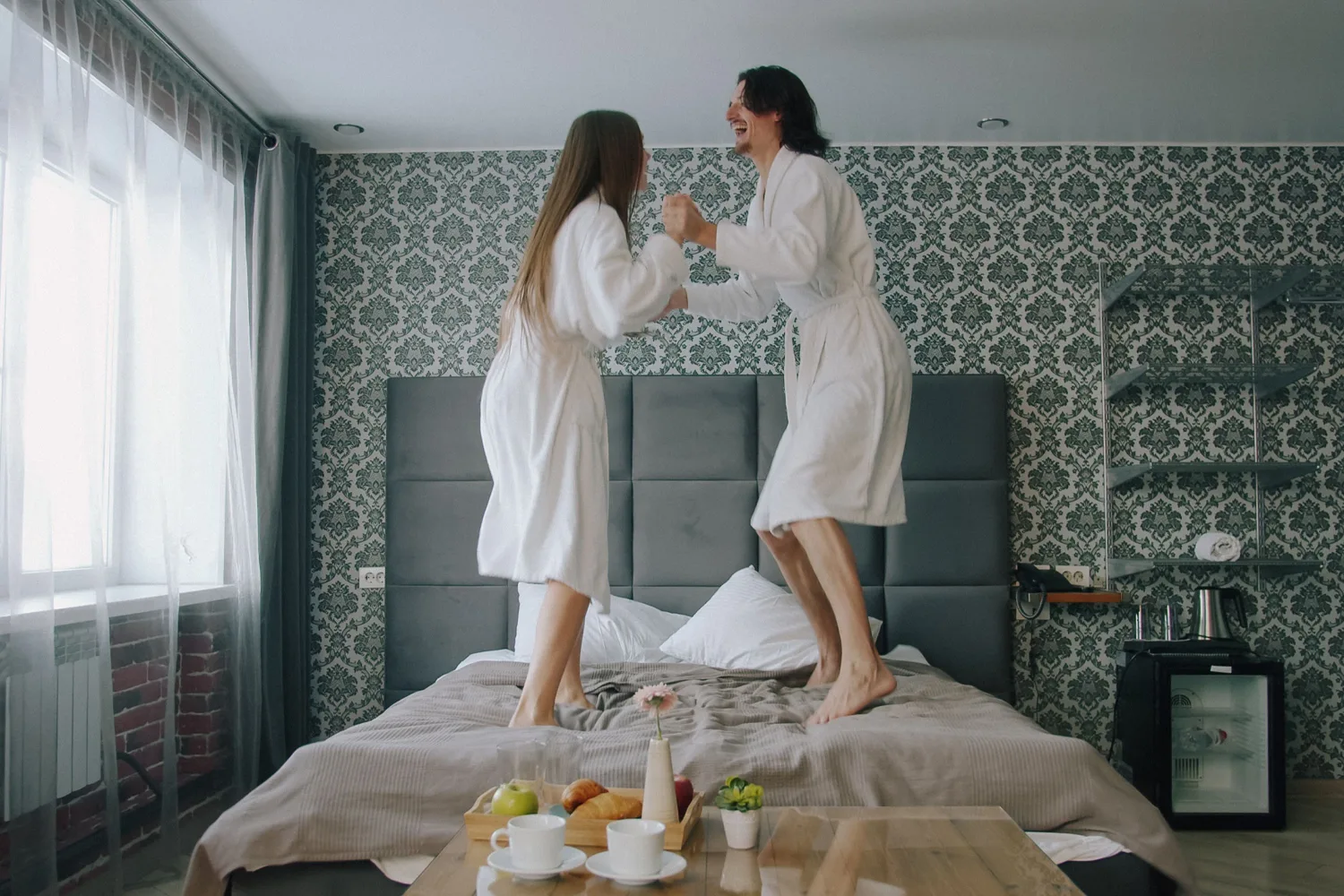 Zwei Personen in Bademänteln springen lachend auf einem Hotelbett, davor ein Tisch mit Frühstück